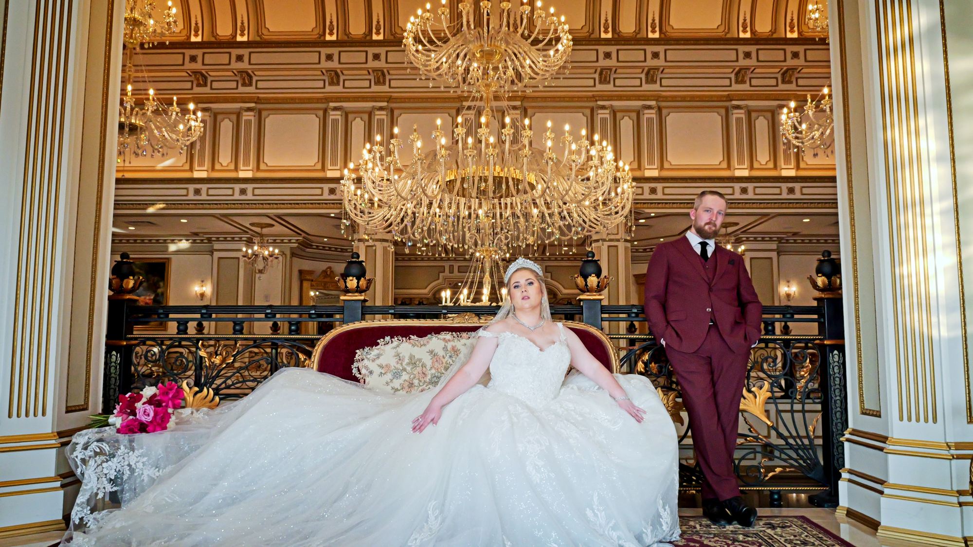 A bride and groom in a grand hotel lobby beneath chandeliers