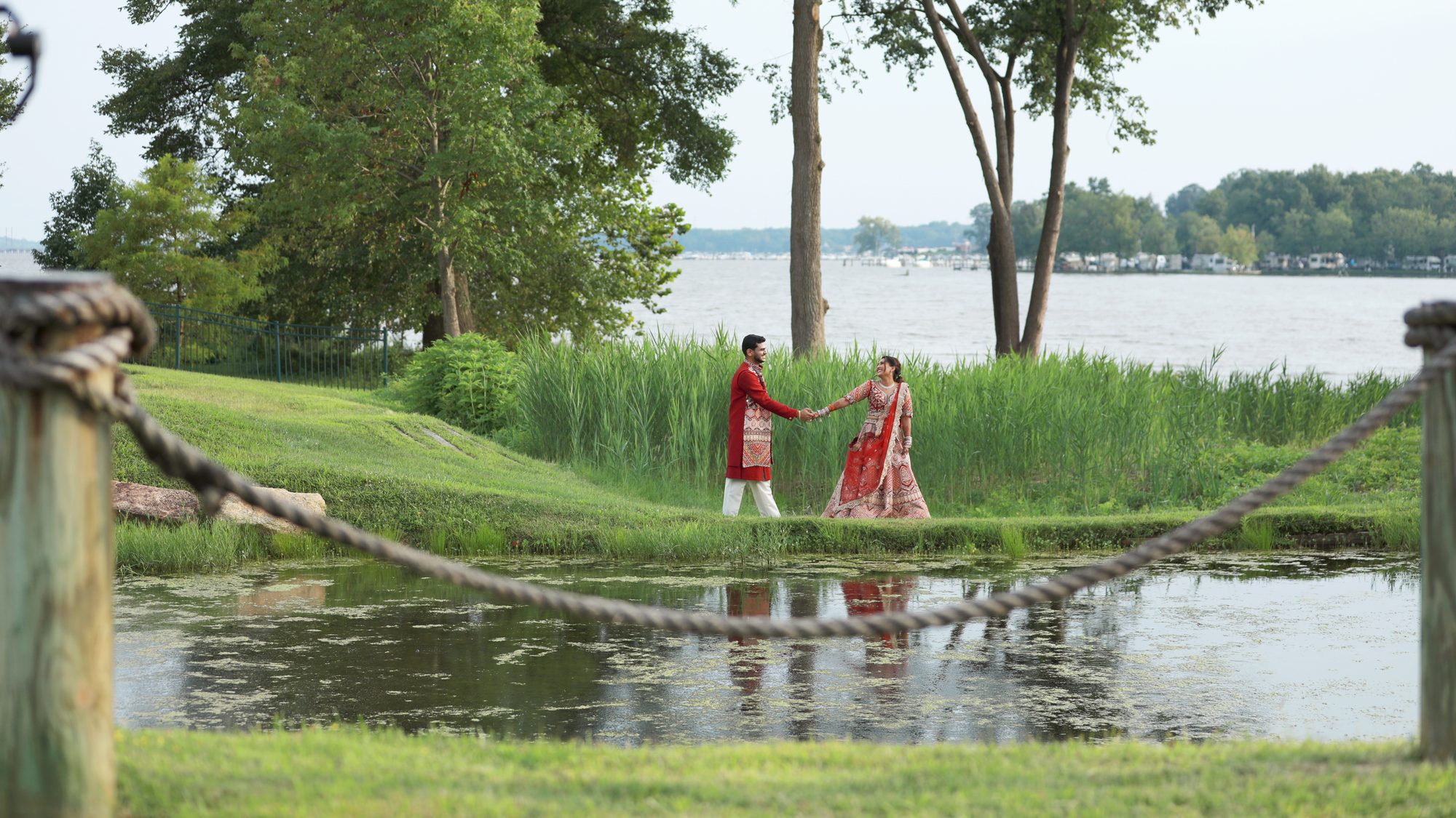 A bride and groom holding hands on a lawn beside a lake