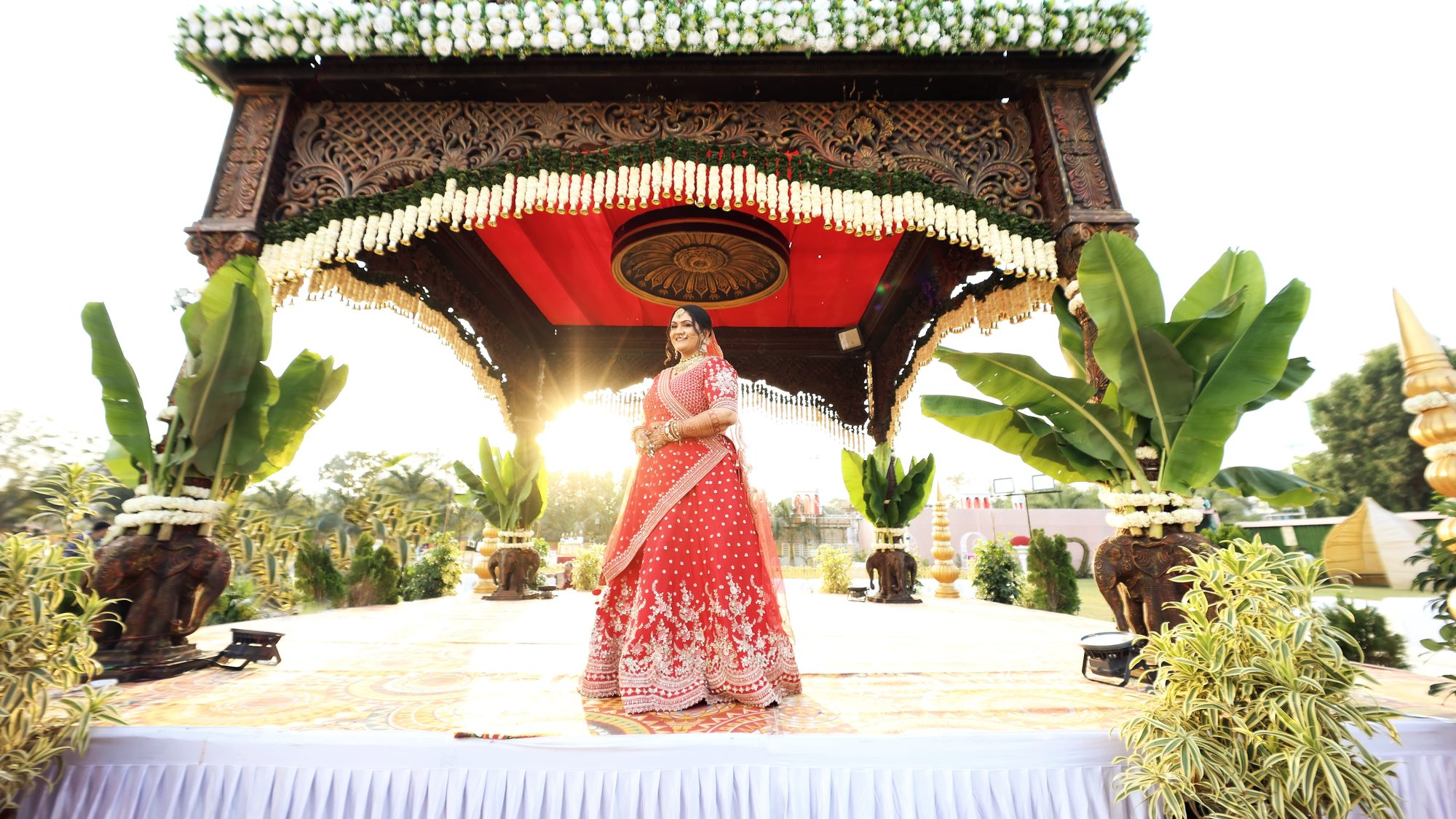 A bride standing beneath a carved wooden mandap in afternoon sun
