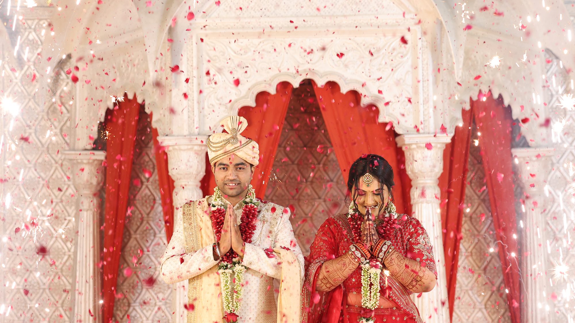 A bride and groom greeting guests with folded hands beneath a floral mandap