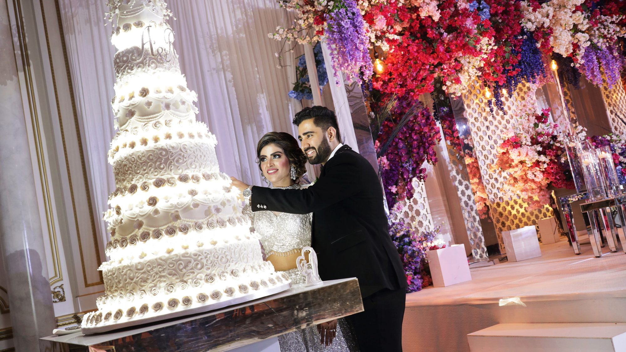 A bride and groom cutting a tall wedding cake beside floral décor